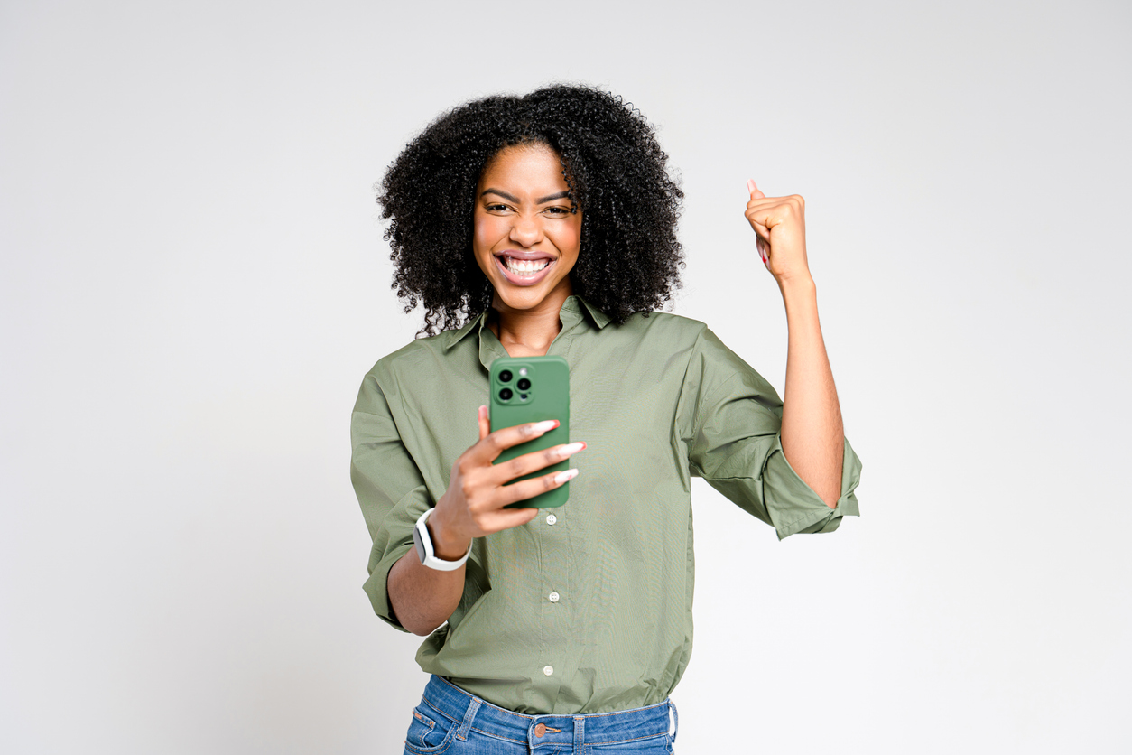 Energetic and triumphant, an African-American woman raises her fists in victory, holding smartphone and receiving good news, embodying success and achievement against a clean background