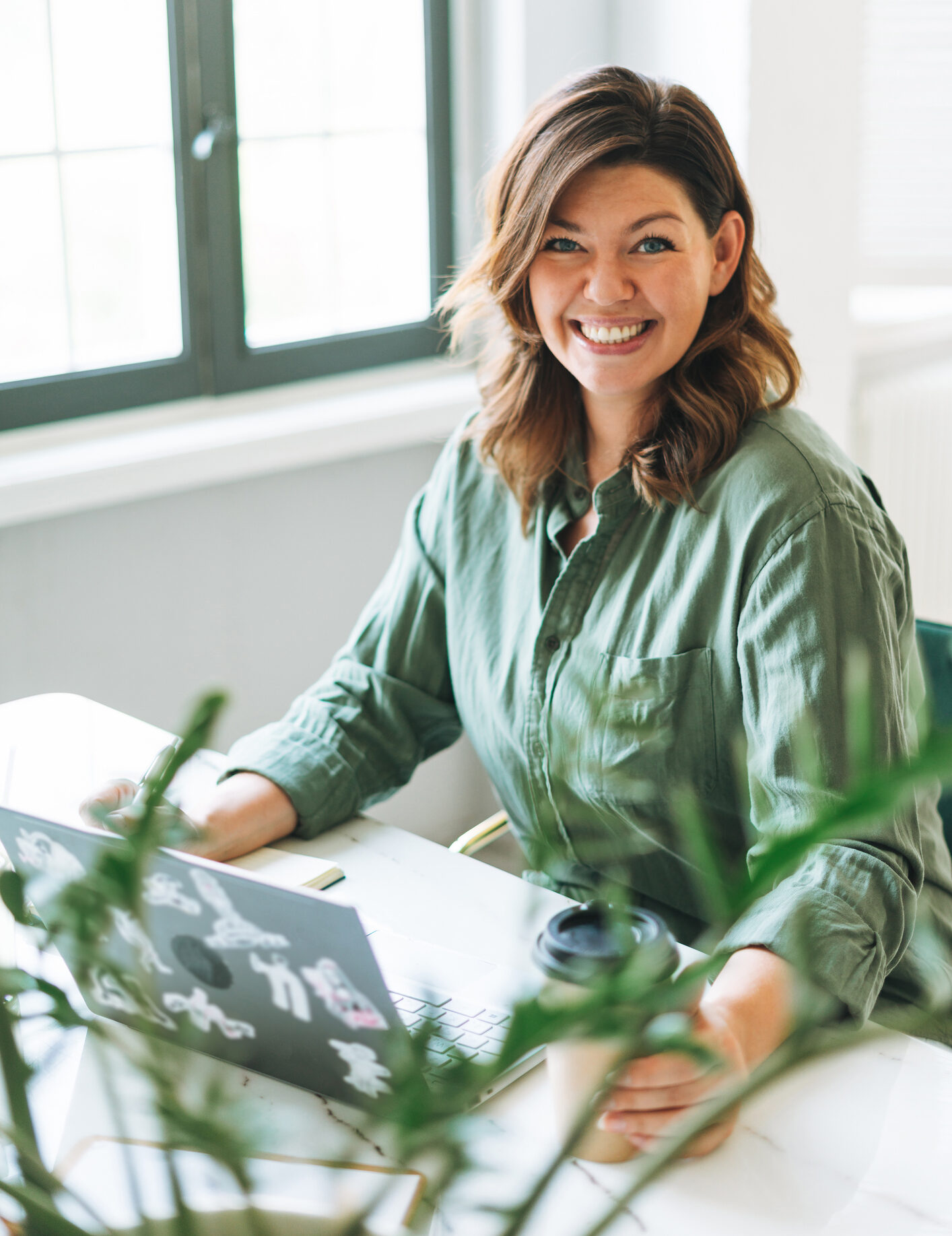 Young smiling brunette woman plus size working at laptop on table with house plant in the bright modern office