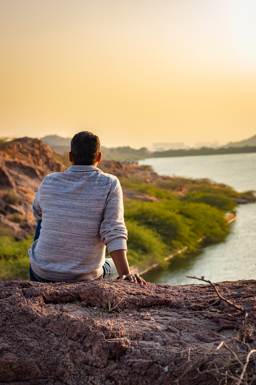 isolated young man sitting at mountain top with lake view from flat angle image is taken at kaylana lake jodhpur rajasthan india.