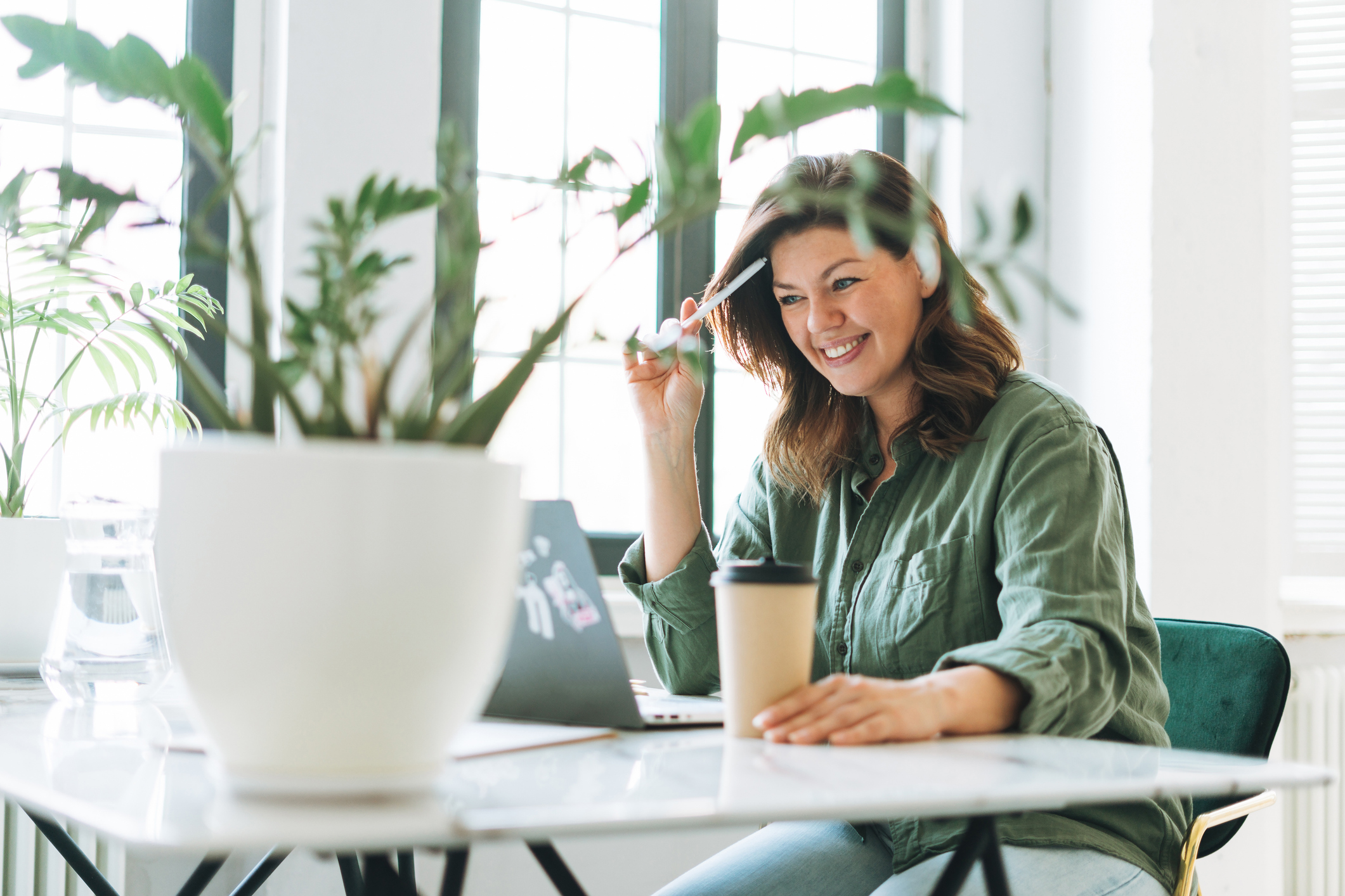 Young smiling brunette woman plus size working at laptop on table with house plant in the bright modern office Young smiling brunette woman plus size working at laptop on table with house plant in the bright modern office
